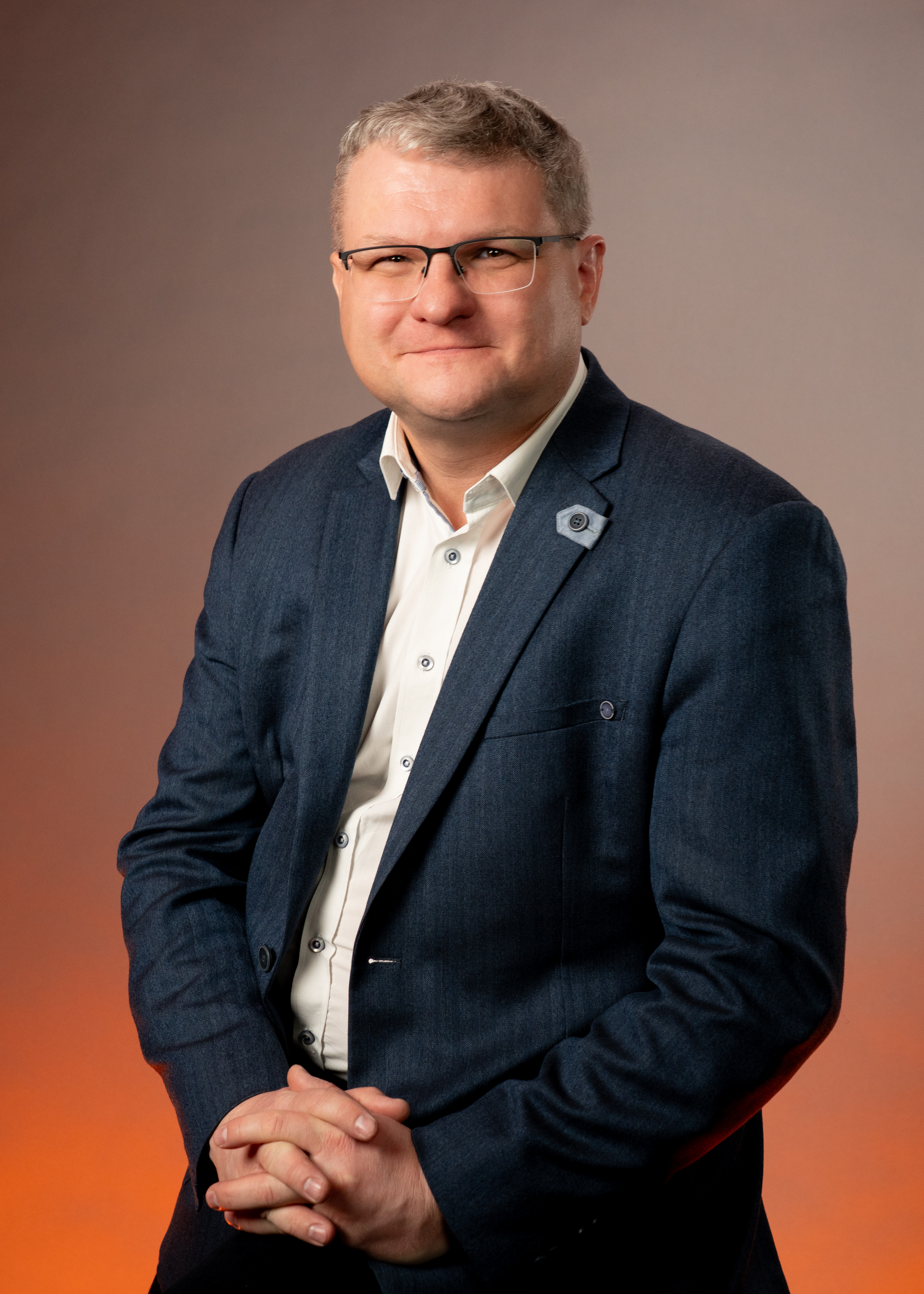 Muž ve středním věku oblečený do tmavého obleku a bílé košile stojí se zkříženými rukama na pozadí s oranžovým gradientem.