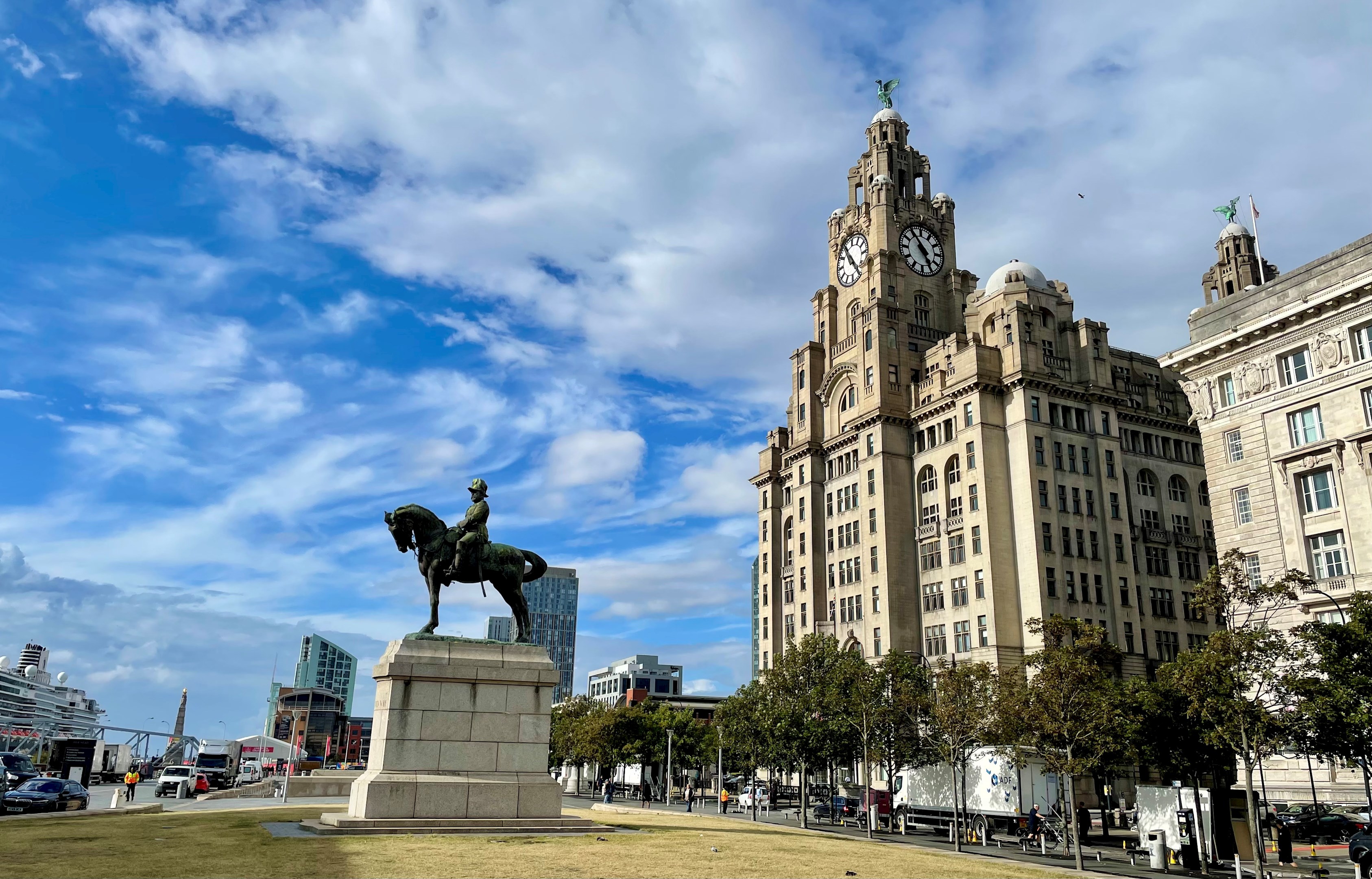 A bronze equestrian statue in the foreground with the iconic Royal Liver Building in the background under a blue sky with scattered clouds in Liverpool.