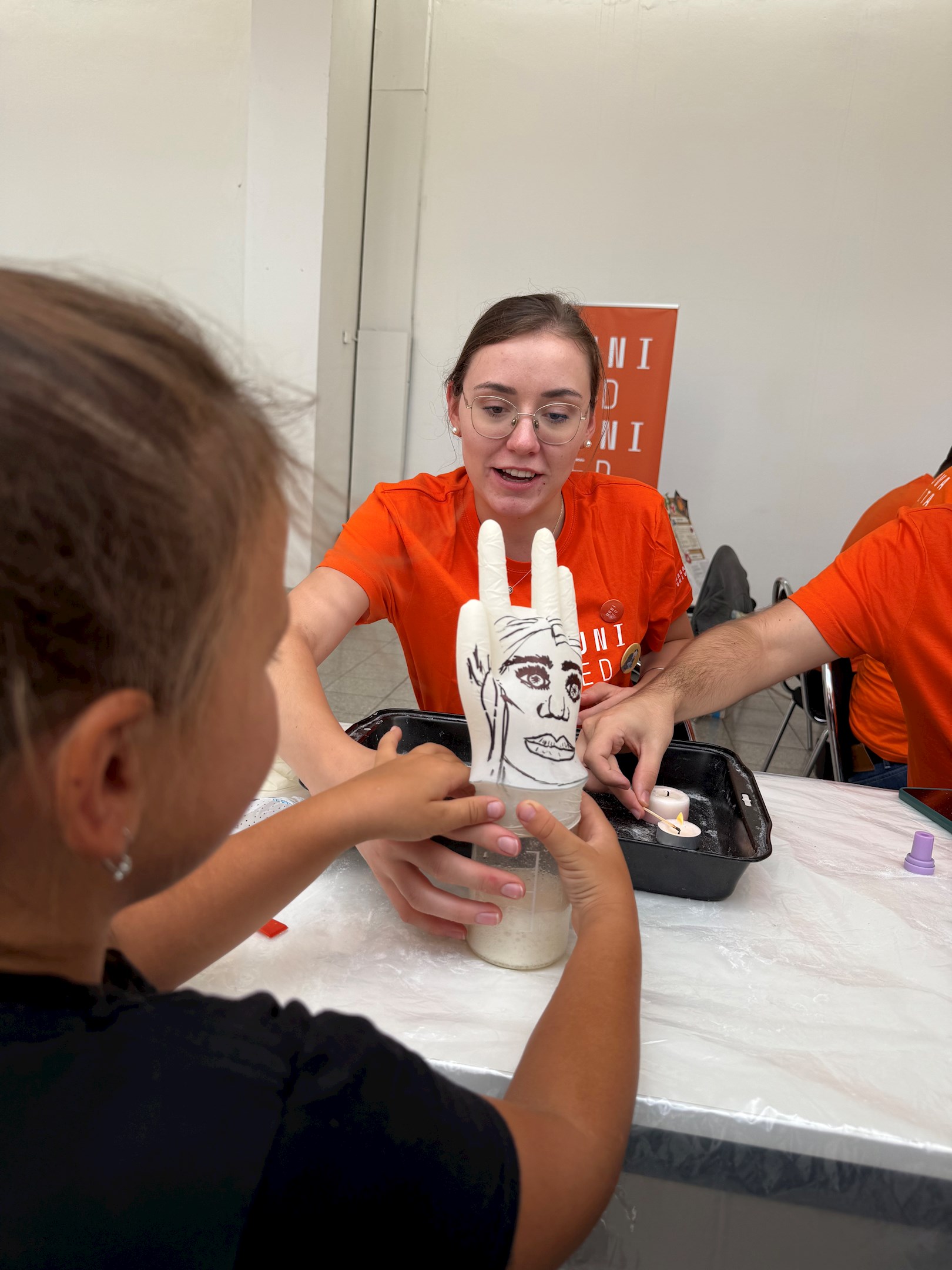 Osoba v oranžovém tričku maluje obličej na ruce dítěte u stolu s kreativními potřebami.