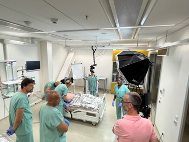 Skupina zdravotnických pracovníků v zelených uniformách stojí kolem lůžka v nemocničním pokoji, kde je umístěn pacient, zatímco filmový štáb je přítomen pro natáčení.